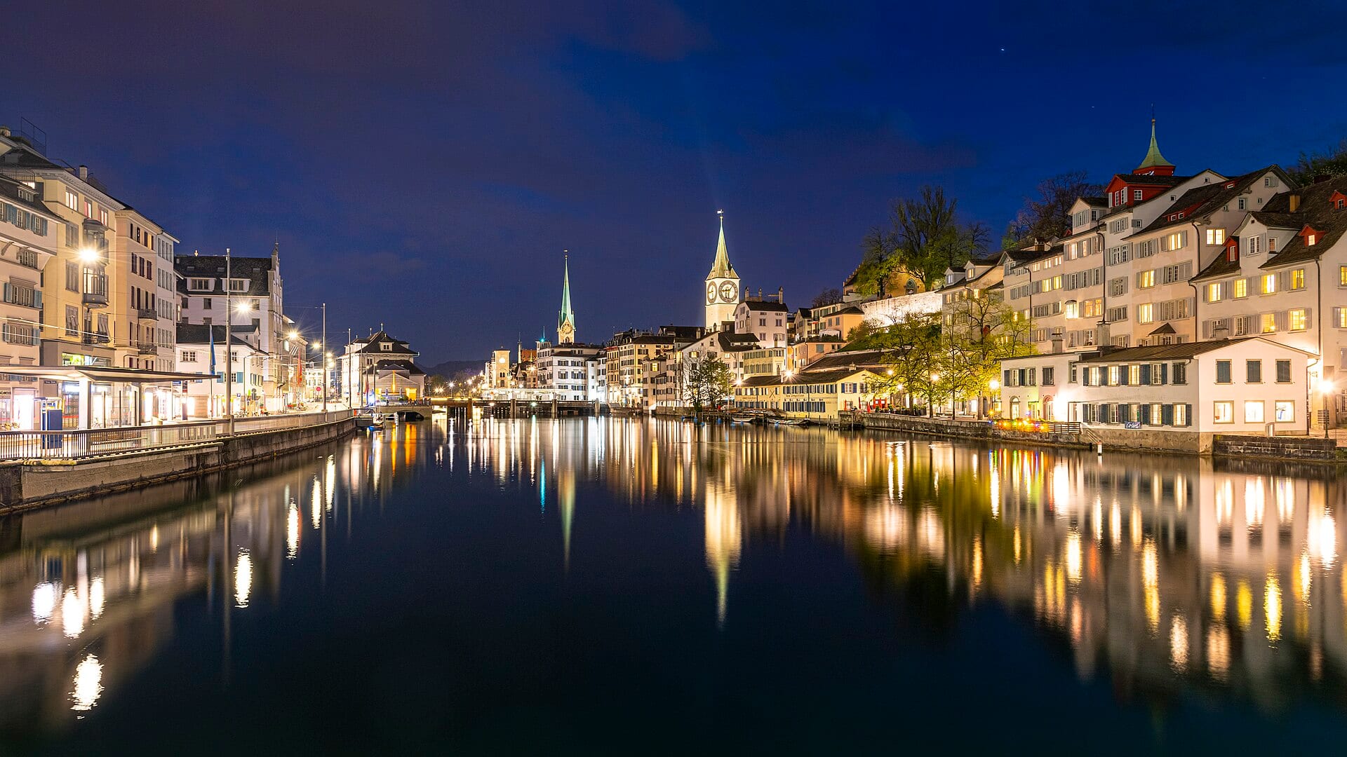 The Limmat in Zurich at dusk, old town houses lit along both banks, a slim church spire and a clock tower behind them, and the lights reflected in still water.