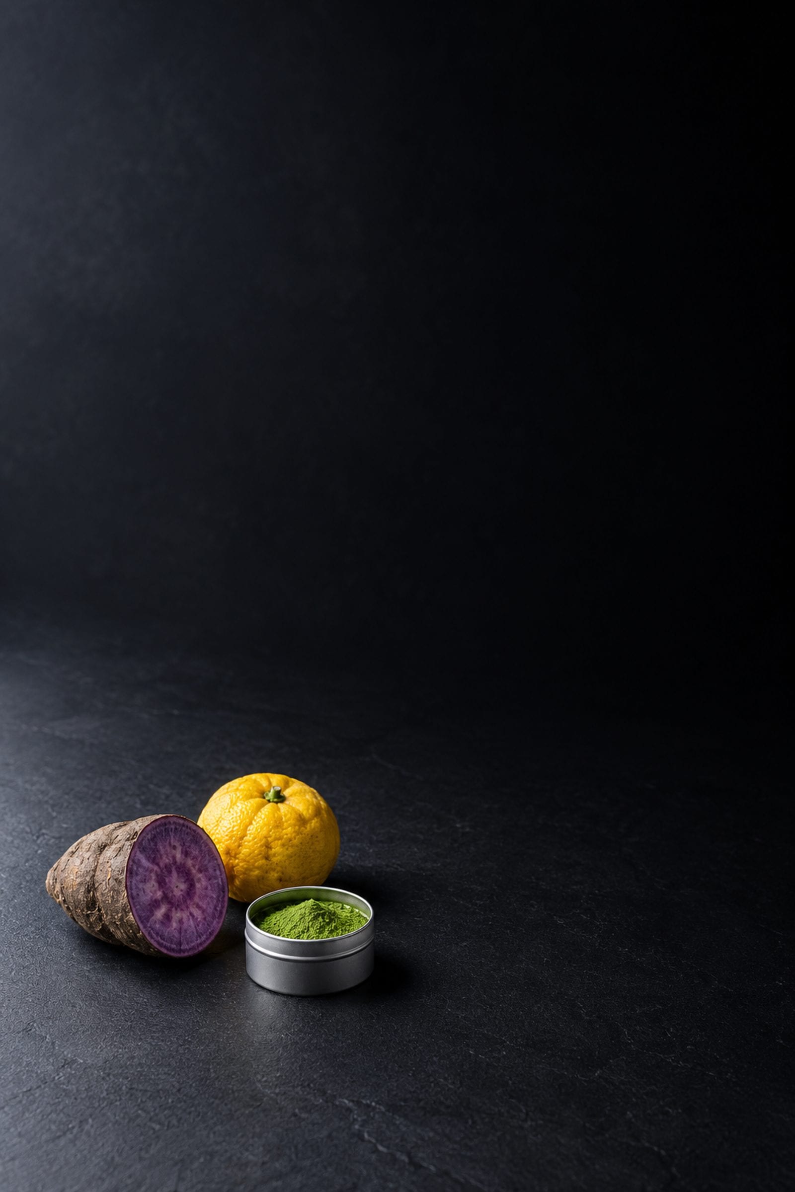 A cut purple ube, a whole yuzu and an open tin of matcha powder, grouped on dark slate with the rest of the frame left empty.