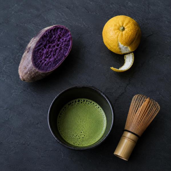 A flat lay of a halved purple ube, a partially peeled yuzu, a bowl of whisked matcha and a bamboo chasen, arranged on dark slate.