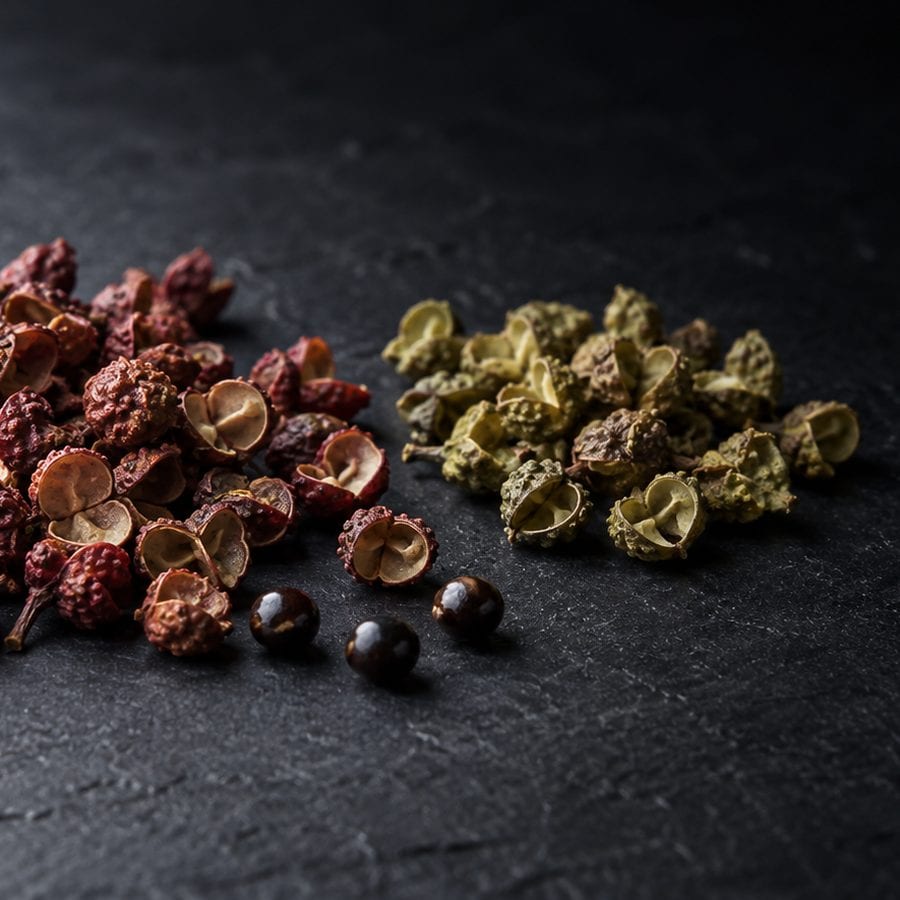 Two heaps of husked peppercorns, red on the left and green on the right, with a few shiny black seeds between them, on dark slate.