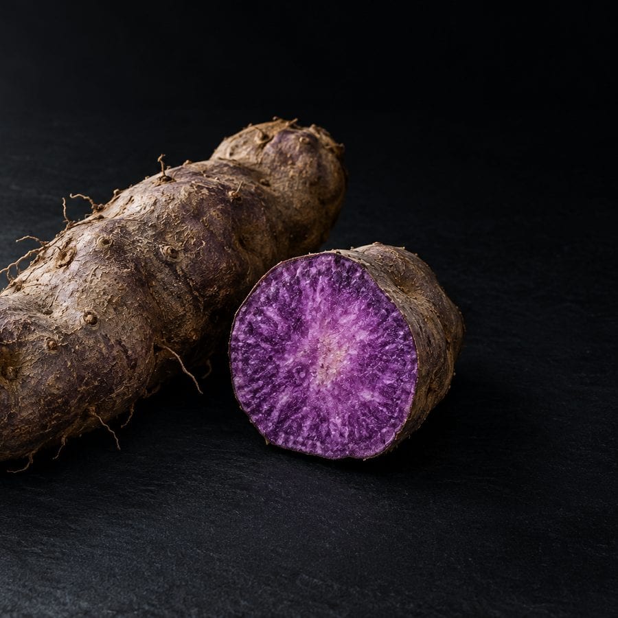A whole purple yam lying beside a cut section, its flesh bright violet with pale marbling, on dark slate.