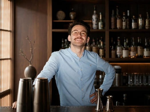 Simon Engel standing behind a bar, hands resting on the counter beside mixing tins and a jigger, bottles on the shelves behind him.